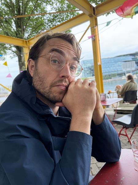 A man with glasses sits thoughtfully at a red table in an outdoor cafe at Commandantsbrug, Marineterrein, Amsterdam